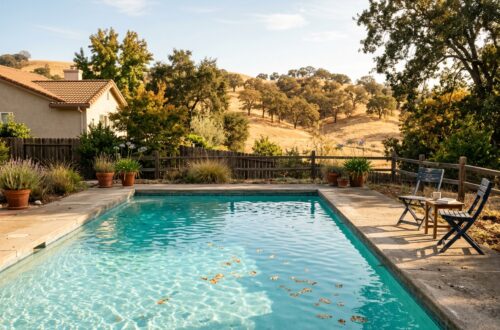 A backyard swimming pool with a house and rolling golden hills beyond, under a clear blue sky.