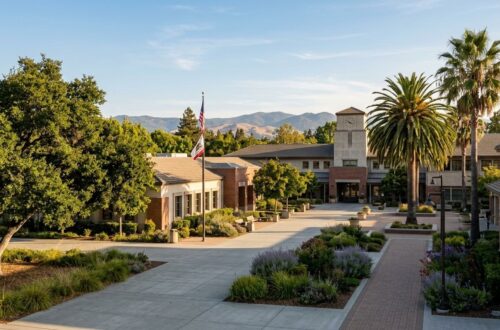 A civic campus with a clock tower, palm trees, and U.S. and California flags under a blue sky.
